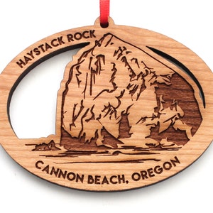May include: Wooden ornament featuring a laser-cut design of Haystack Rock at Cannon Beach, Oregon. The oval-shaped ornament has a red ribbon for hanging. The text "HAYSTACK ROCK" and "CANNON BEACH, OREGON" are carved into the wood.