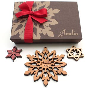 Op de afbeelding: Een set van drie houten sneeuwvlokversieringen in een bruine cadeaudoos met een rode strik. De versieringen zijn gepersonaliseerd met de naam "Amelia" en het jaar "2017".
