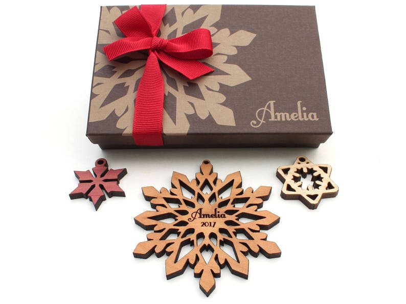Op de afbeelding: Een set van drie houten sneeuwvlokversieringen in een bruine cadeaudoos met een rode strik. De versieringen zijn gepersonaliseerd met de naam "Amelia" en het jaar "2017".