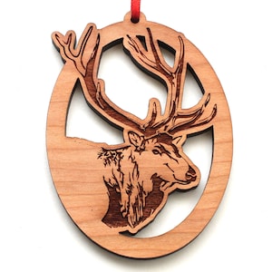 May include: A wooden Christmas ornament featuring a detailed laser-cut design of an elk's head and antlers within an oval frame. The ornament is made of light brown wood and has a red ribbon for hanging.