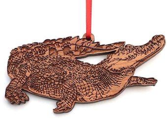 May include: A wooden ornament of a brown alligator with a detailed texture. The ornament is hanging from a red ribbon.