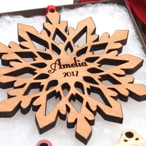 Op de afbeelding: Een houten sneeuwvlok ornament met een bruine vlek en een lasergegraveerd ontwerp. De ornament is gepersonaliseerd met de naam "Amelia" en het jaar "2017".