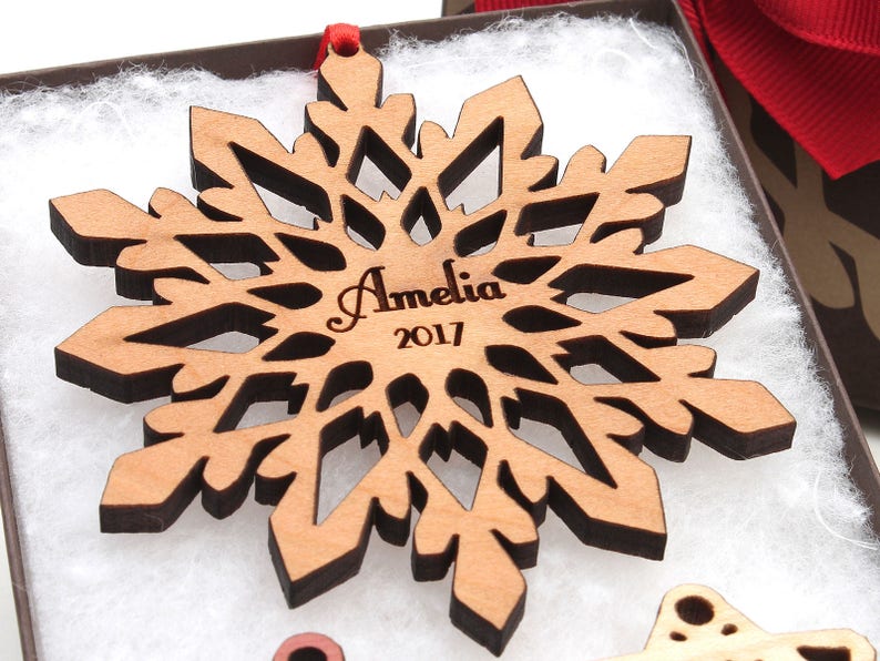 Op de afbeelding: Een houten sneeuwvlok ornament met een bruine vlek en een lasergegraveerd ontwerp. De ornament is gepersonaliseerd met de naam "Amelia" en het jaar "2017".