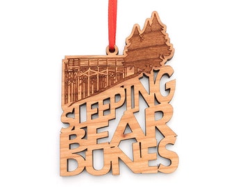 Puede incluir: Adorno de madera con un diseño de corte láser de Sleeping Bear Dunes National Lakeshore, Michigan. El adorno presenta una silueta de las dunas de Sleeping Bear con las palabras "Sleeping Bear Dunes" cortadas en la madera.