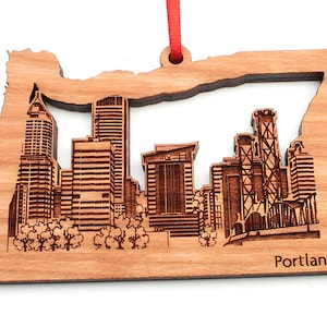 May include: Wooden ornament shaped like the state of Oregon with a laser-engraved cityscape of Portland, Oregon. The ornament is brown and has a red ribbon attached.
