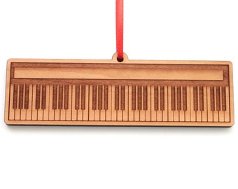Pode incluir: Ornamento de madeira em forma de teclado de piano com uma fita vermelha presa. O ornamento é feito de madeira marrom claro e tem um design detalhado das teclas do piano.