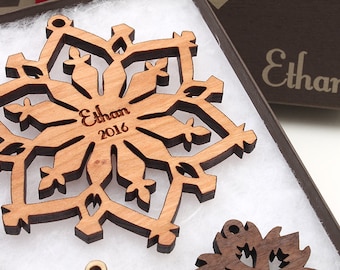 Op de afbeelding: Een houten sneeuwvlok ornament met de naam "Ethan" en het jaar "2016" erop gegraveerd. Het ornament zit in een doos met de naam "Ethan" op de zijkant.