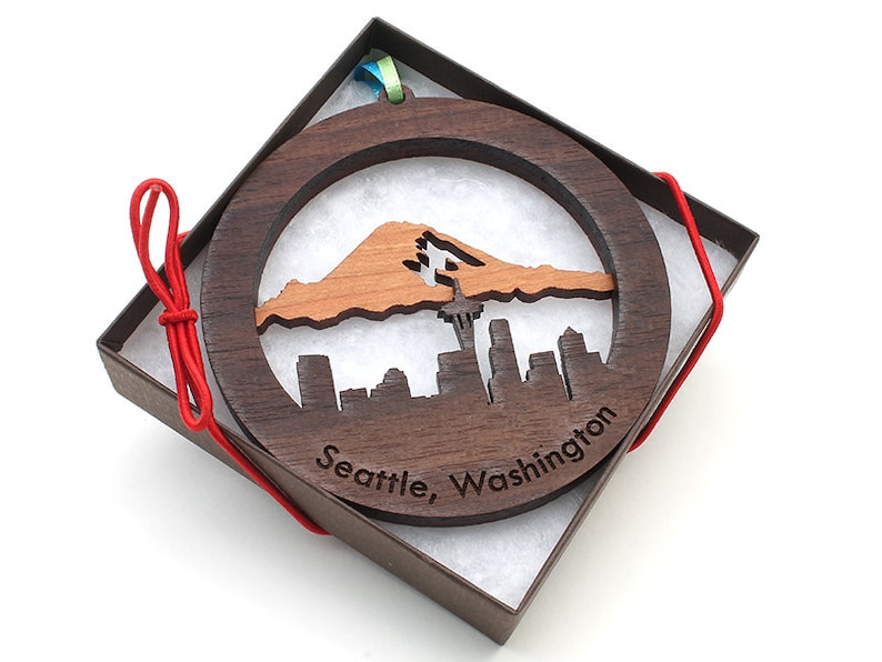 May include: A round wooden ornament with a cutout design of the Seattle skyline and Mount Rainier. The ornament is in a brown box with red ribbon.