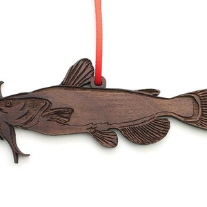 May include: A wooden ornament shaped like a catfish with a red ribbon attached. The ornament is brown and has a detailed carving of the fish.