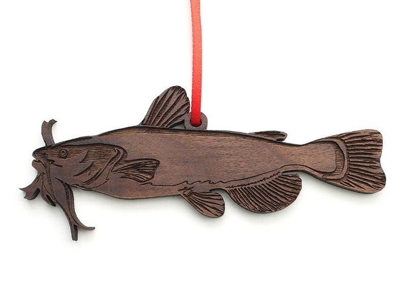 May include: A wooden ornament shaped like a catfish with a red ribbon attached. The ornament is brown and has a detailed carving of the fish.
