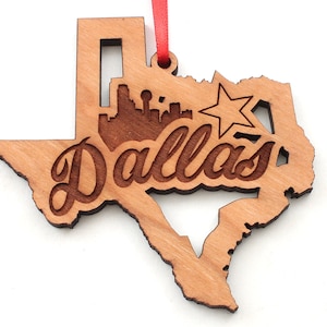 May include: A wooden ornament in the shape of Texas, featuring the city of Dallas. The ornament has a cutout of the Dallas skyline and a star. The word "Dallas" is written in a cursive font. A red ribbon is attached for hanging.