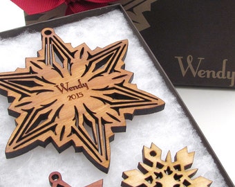 Op de afbeelding: Een houten sneeuwvlok ornament met een gepersonaliseerde inscriptie die "Wendy 2015" luidt. Het ornament bevindt zich in een bruine doos met wit tissuepapier.