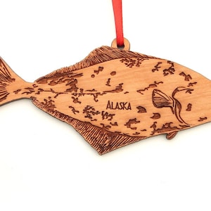 May include: A wooden ornament shaped like a halibut fish with the word "ALASKA" engraved on it. The ornament is a light brown colour and has a detailed design.