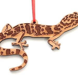 Peut inclure: Une décoration en bois en forme de gecko avec des marques brunes et noires. La décoration est suspendue à un ruban rouge.