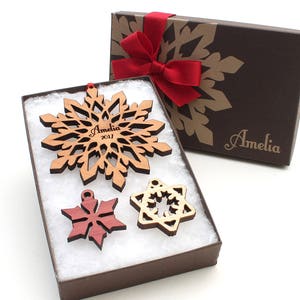 Op de afbeelding: Een set van drie houten sneeuwvlokversieringen in een bruine geschenkdoos met een rood lint. De grootste sneeuwvlok is gepersonaliseerd met de naam "Amelia" en het jaar "2017".