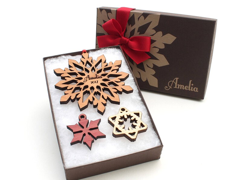 Op de afbeelding: Een set van drie houten sneeuwvlokversieringen in een bruine geschenkdoos met een rood lint. De grootste sneeuwvlok is gepersonaliseerd met de naam "Amelia" en het jaar "2017".