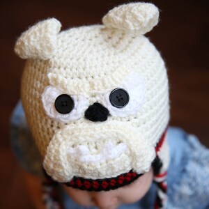 May include: A cream-colored crocheted bulldog hat with black button eyes and a black nose. The hat has small, upright ears and a white muzzle. Braided ties with red and black accents hang from the hat.