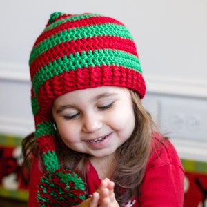 May include: A young child wearing a red and green striped crocheted elf hat with a pom-pom. The child is wearing a red long-sleeved shirt and is holding their hands together in front of them.