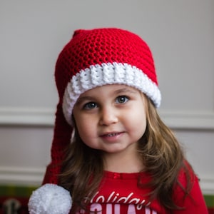 May include: A young girl wearing a red and white crocheted Santa hat with a white pom-pom, a red long-sleeved shirt with the text "Holiday Squad" and a pink graphic, and pink and white pajamas.