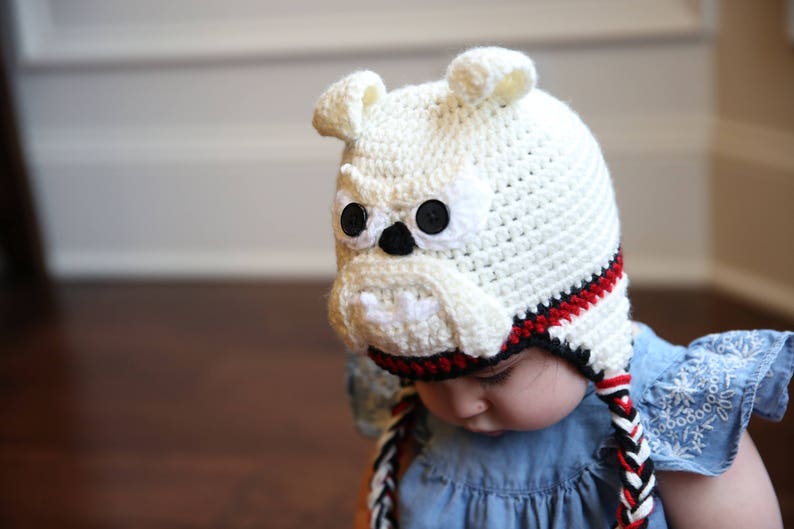 May include: A handmade, off-white crochet hat shaped like a bulldog's head, featuring black button eyes, a black nose, and a white muzzle. The hat has red and black trim and braided ties.