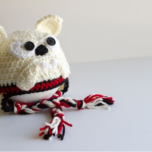 May include: A handmade crochet owl with button eyes and a black beak. The owl is cream-colored with a red, black, and white striped band around its middle. A matching scarf is draped in front.