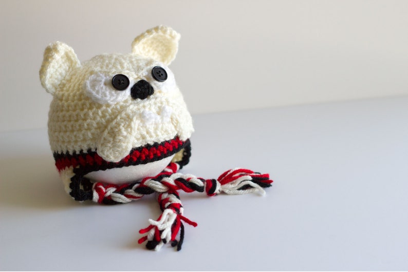 May include: A handmade crochet owl with button eyes and a black beak. The owl is cream-colored with a red, black, and white striped band around its middle. A matching scarf is draped in front.