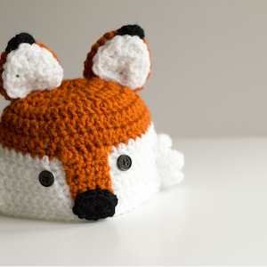May include: A crocheted fox hat with white and orange yarn. The hat has black button eyes and a black nose.