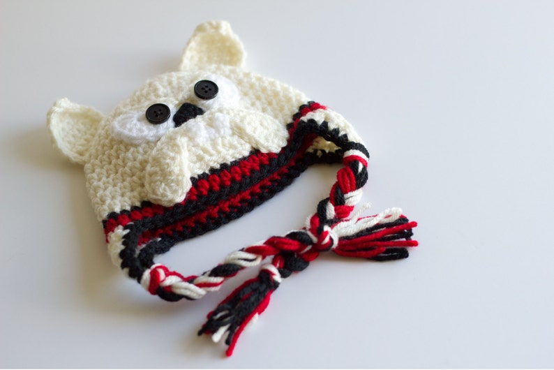 May include: A cream-colored crocheted owl hat with black button eyes and ear details. The hat features red, black, and white trim and a braided tassel. A whimsical and cozy accessory.