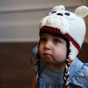 May include: A white crocheted hat with a bulldog face design, featuring black button eyes and small ears. The hat has a black and red striped band and braided ties. The child is wearing a blue denim dress.