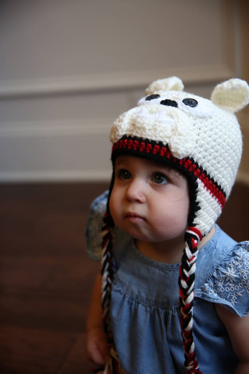 May include: A white crocheted hat with a bulldog face design, featuring black button eyes and small ears. The hat has a black and red striped band and braided ties. The child is wearing a blue denim dress.