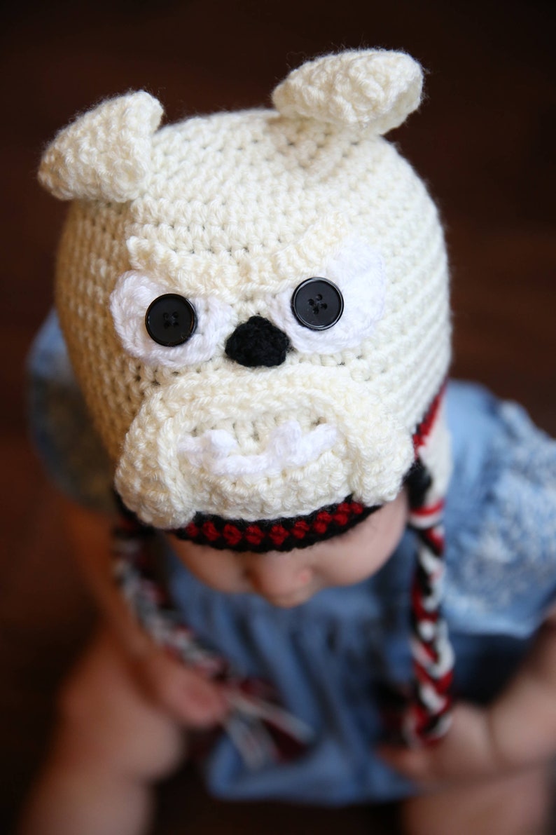 May include: A cream-colored crocheted bulldog hat with black button eyes and a black nose. The hat has small, upright ears and a white muzzle. Braided ties with red and black accents hang from the hat.