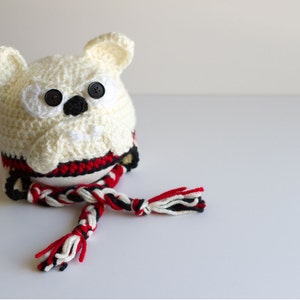 May include: A handmade, crocheted owl-shaped hat in cream, with black button eyes and a black nose. The hat features a red, black, and white striped band and a matching braided tassel. The owl has small, cream-colored ears.