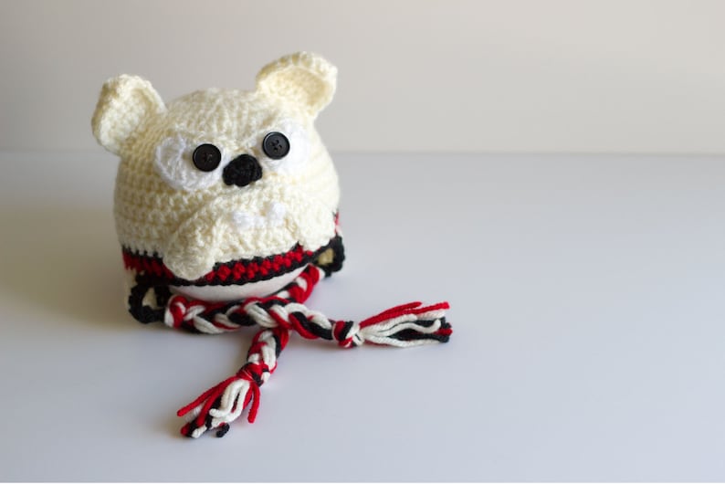 May include: A handmade, crocheted owl-shaped hat in cream, with black button eyes and a black nose. The hat features a red, black, and white striped band and a matching braided tassel. The owl has small, cream-colored ears.