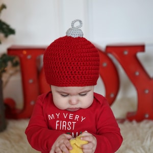 May include: A baby wearing a red crocheted ornament hat with a silver top, sitting in front of a red JOY sign. The baby is holding a yellow toy in their hands.
