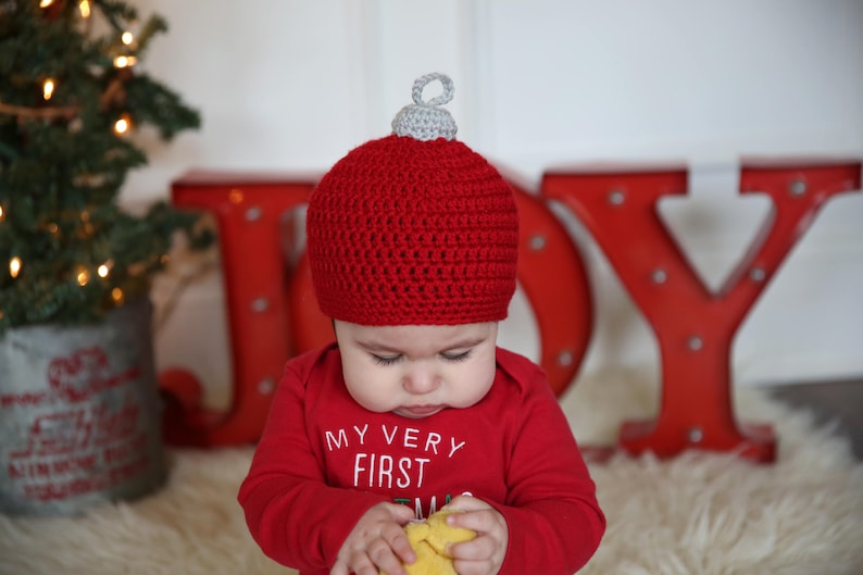 May include: A baby wearing a red crocheted ornament hat with a silver top, sitting in front of a red JOY sign. The baby is holding a yellow toy in their hands.