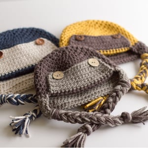 May include: Three crocheted hats with braided tails and wooden buttons. The hats are in shades of brown, blue, and yellow.
