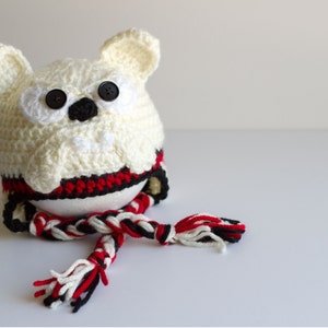 May include: A handmade, off-white crocheted owl-shaped hat with black button eyes and a black nose. The hat has red and black accents around the base and a matching scarf. The owl has large, rounded ears.