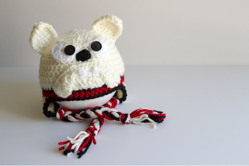 May include: A handmade, off-white crocheted owl-shaped hat with black button eyes and a black nose. The hat has red and black accents around the base and a matching scarf. The owl has large, rounded ears.