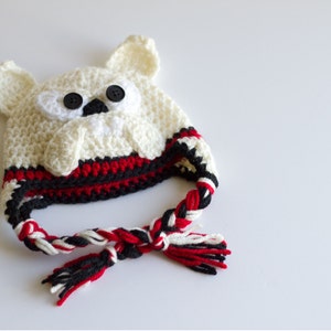 May include: A handmade crochet hat in the shape of a polar bear. The hat is primarily off-white with black button eyes and a black nose. The brim features red and black stripes, and the braided ties are red, black, and white.