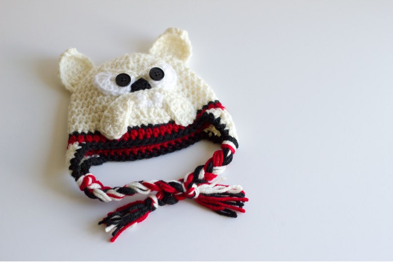 May include: A handmade crochet hat in the shape of a polar bear. The hat is primarily off-white with black button eyes and a black nose. The brim features red and black stripes, and the braided ties are red, black, and white.