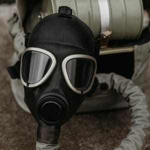 May include: A black gas mask with silver-edged lenses and a circular filter. The mask is connected to a grey hose and rests in a matching grey canvas bag. The filter is a light olive green colour.
