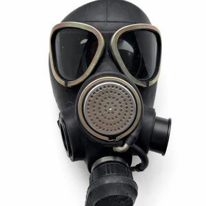 Puede incluir: Máscara de gas negra con lentes grandes y oscuras y un filtro circular. La máscara tiene una textura engomada y una serie de accesorios, incluido un tubo respiratorio. Las lentes están enmarcadas con un borde metálico.