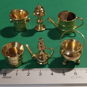 May include: Six miniature gold-toned metal objects, including a watering can, a bucket, a lamp, a pitcher, and two bowls, are arranged on a green surface. A ruler is placed below the objects, with the numbers 0 to 6 marked in centimeters.