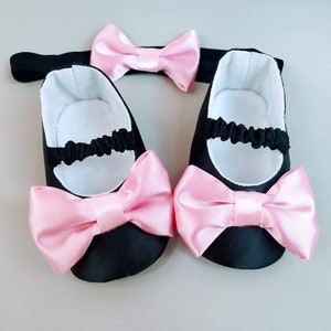 Peut inclure: Un ensemble de chaussures de b&eacute;b&eacute; noires avec des n&oelig;uds en satin rose et un bandeau assorti avec un n&oelig;ud en satin rose.