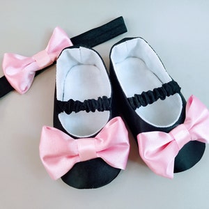 Peut inclure: Une paire de chaussures de b&eacute;b&eacute; noires avec des n&oelig;uds en satin rose et un bandeau noir avec un n&oelig;ud en satin rose.