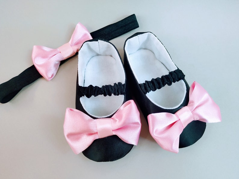 Peut inclure: Une paire de chaussures de b&eacute;b&eacute; noires avec des n&oelig;uds en satin rose et un bandeau noir avec un n&oelig;ud en satin rose.