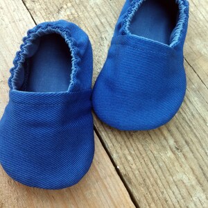 Op de afbeelding: Een paar felblauwe babyschoentjes gemaakt van een textiel met structuur. De schoentjes hebben een ronde neus en elastiek rond de opening. Ze liggen op een rustiek houten oppervlak.