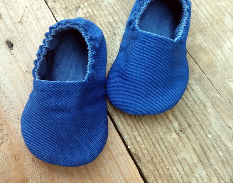 Op de afbeelding: Een paar felblauwe babyschoentjes gemaakt van een textiel met structuur. De schoentjes hebben een ronde neus en elastiek rond de opening. Ze liggen op een rustiek houten oppervlak.