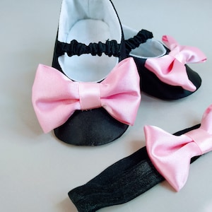 Peut inclure: Une paire de chaussures de b&eacute;b&eacute; noires avec des n&oelig;uds en satin rose et un bandeau noir avec un n&oelig;ud en satin rose.