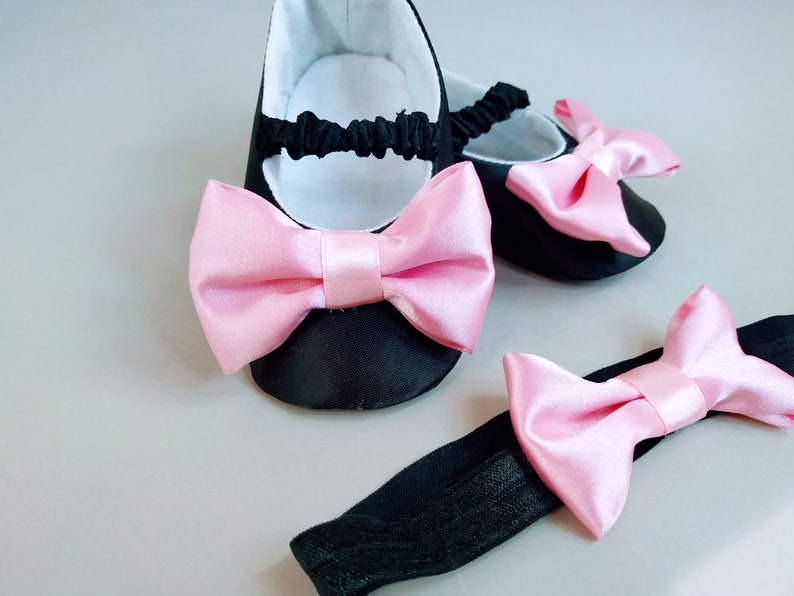 Peut inclure: Une paire de chaussures de b&eacute;b&eacute; noires avec des n&oelig;uds en satin rose et un bandeau noir avec un n&oelig;ud en satin rose.